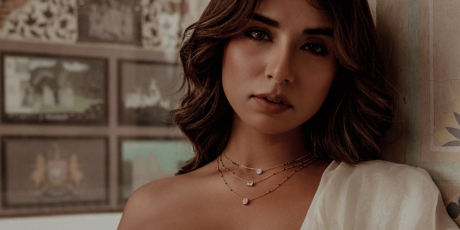 Beautiful South Asian woman wearing a modern mangal trendy modern mangalsutra featuring triple-layered gold chains with delicate black beads and elegant square-cut diamond pendants. She is dressed in a creamy ivory off-shoulder outfit with subtle pleating. The background showcases a vintage mural wall with traditional artwork in muted tones, enhancing the regal, timeless ambiance. The model gazes confidently at the camera from a slight side angle, highlighting the feminine and contemporary essence of the mo