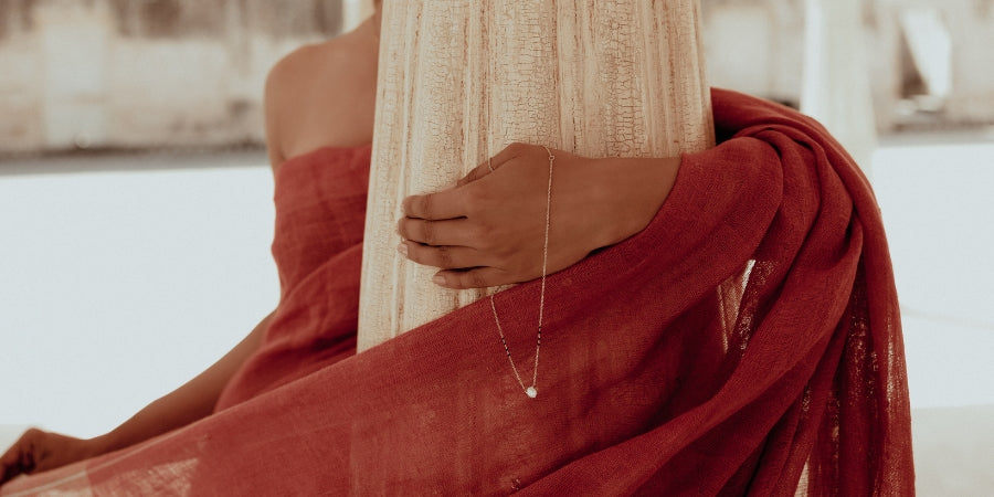 Beautiful South Asian woman wearing a modern mangal trendy modern mangalsutra hand chain featuring a delicate gold chain with a single diamond pendant extending from the wrist to the finger. Draped in a deep red muslin saree with soft golden undertones, she gracefully embraces a carved sandstone pillar. The natural daylight and minimalistic courtyard background accentuate the serene elegance and timeless beauty of modern mangal's contemporary mangalsutra design.