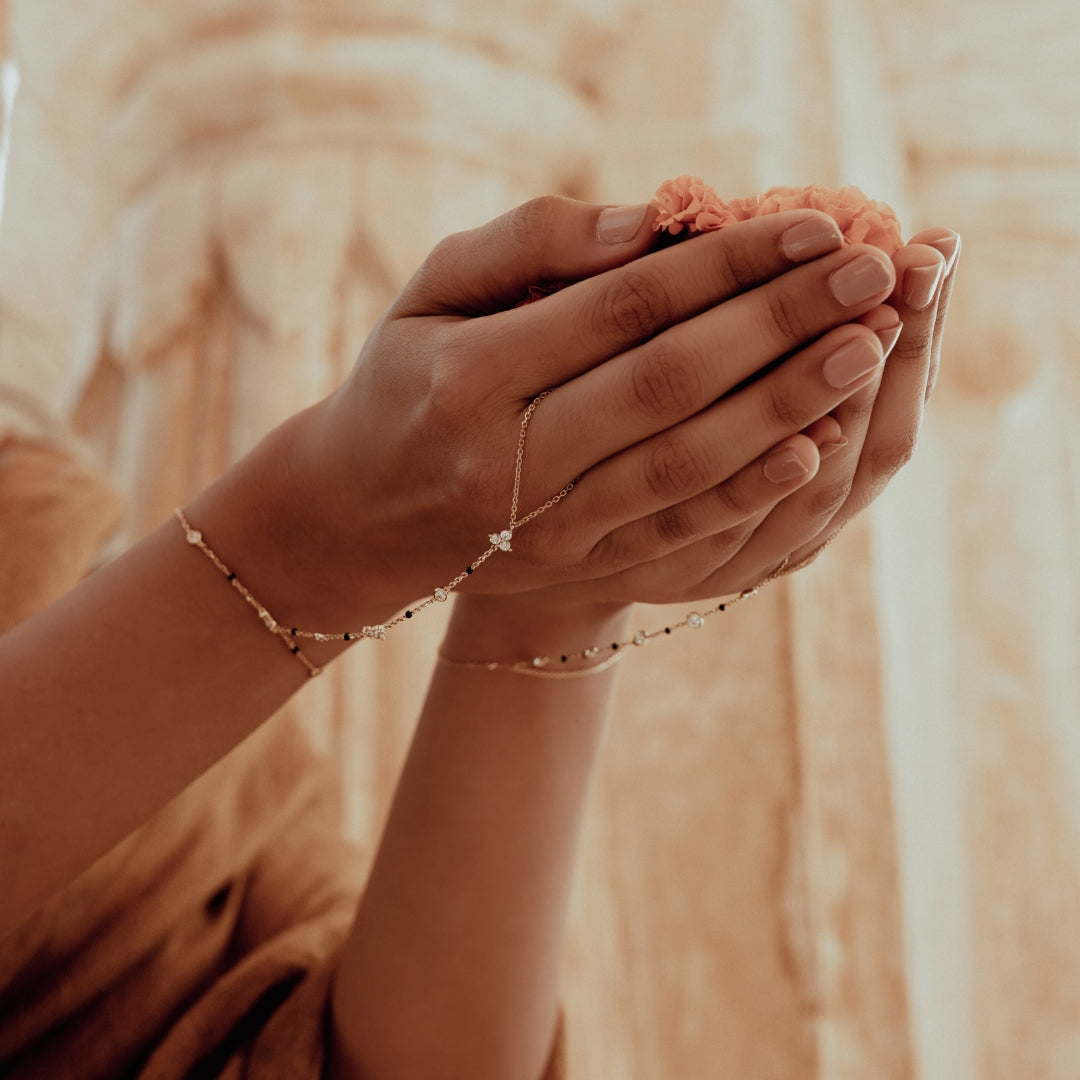 Modern mangal handchain mangalsutra on South Asian woman holding coral flowers, wearing beige saree in soft sandstone temple backdrop.