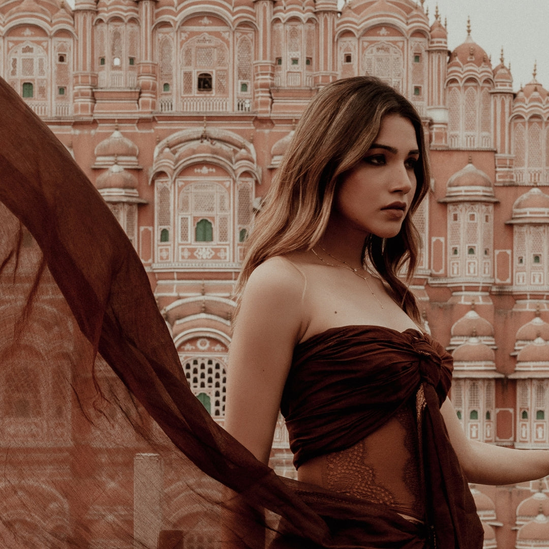 Beautiful South Asian woman wearing a Modern Mangal trendy modern mangalsutra featuring a delicate layered gold chain and diamond pendant. She is styled in a rich maroon draped outfit with sheer lace detailing, standing in front of the iconic terracotta Hawa Mahal façade in Jaipur. The model is captured in a side profile angle, with her dupatta flowing, creating a dramatic yet elegant composition that highlights the contemporary craftsmanship of the mangalsutra by Modern Mangal.