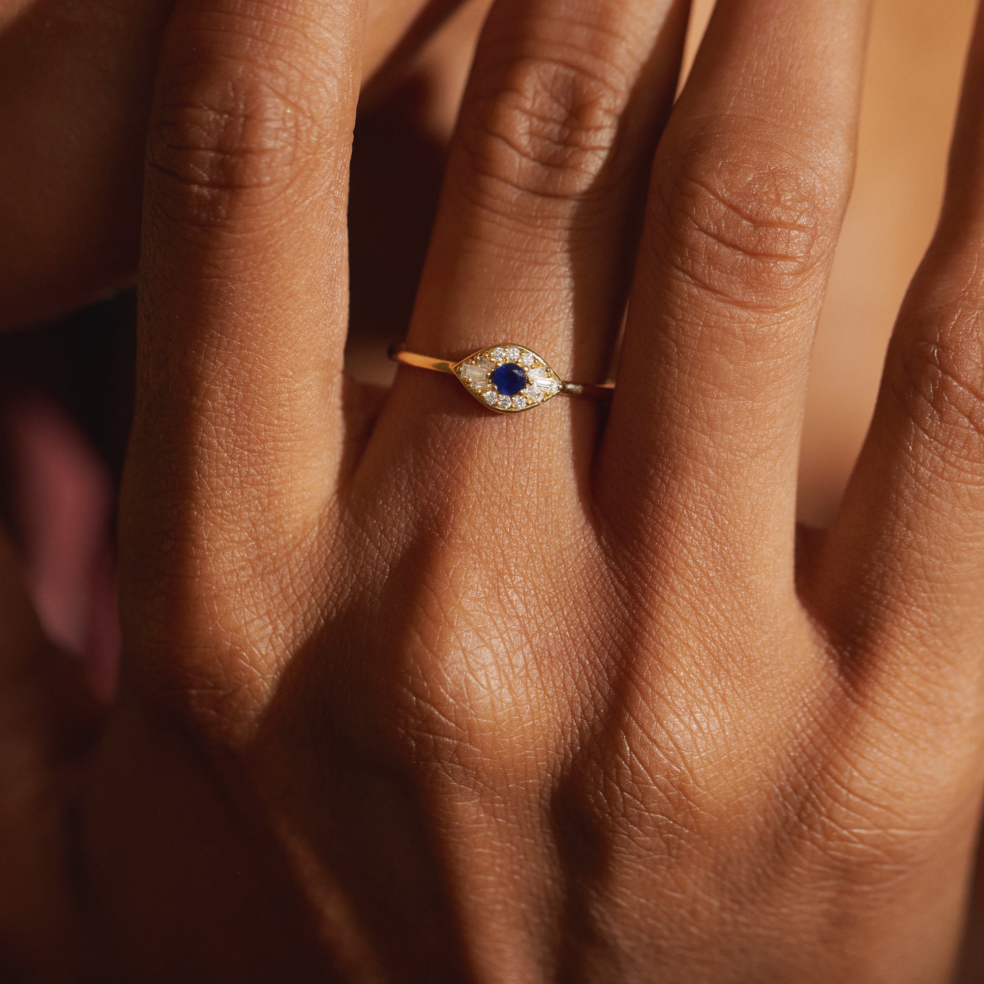 Detailed hand view of the Modern Mangal Zuri Ring in 18K yellow gold, featuring a protective evil eye silhouette centered with a blue sapphire and bordered by precision-set diamonds, embodying refined symbolism and bold design.