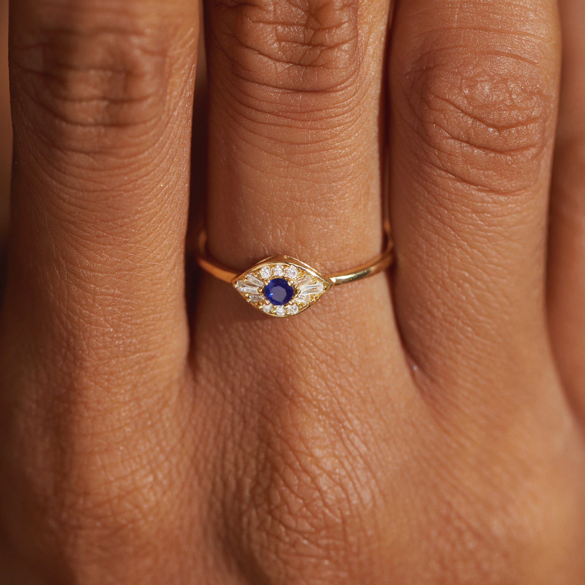 Close-up of the Modern Mangal Zuri Ring worn on a hand, showcasing the marquise-shaped evil eye design with a vivid blue sapphire center surrounded by round and baguette diamonds, all set in 18K yellow gold.
