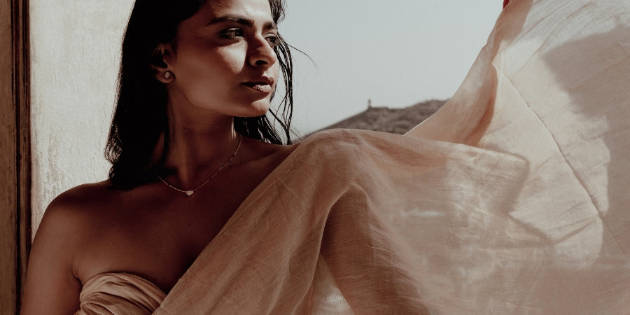 Beautiful South Asian woman wearing a modern mangal trendy modern mangalsutra design featuring a round-cut diamond pendant set on a dainty gold chain with traditional black bead accents. She is gracefully framed against a sunlit rustic window with a soft breeze catching the sheer beige fabric of her sari, styled in an elegant off-shoulder wrap. The photo captures a side-angle view with soft natural light that enhances the timeless charm of modern mangal's fine jewelry.
