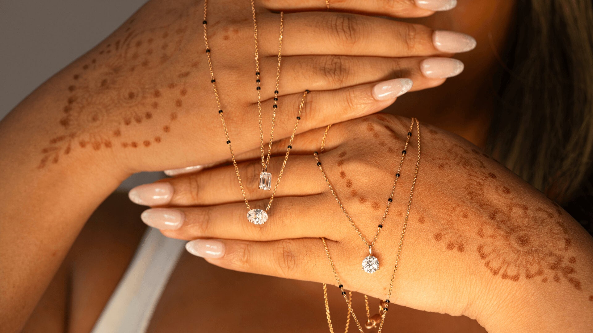 Hands adorned with henna designs, showcasing multiple gold mangalsutras with sparkling diamond pendants.