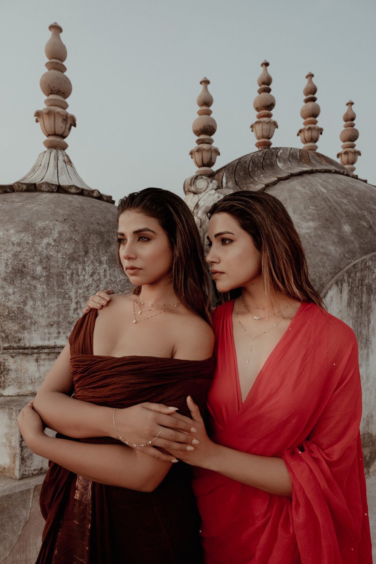Two women in red and maroon drapes holding each other beside sandstone domes in Jaipur