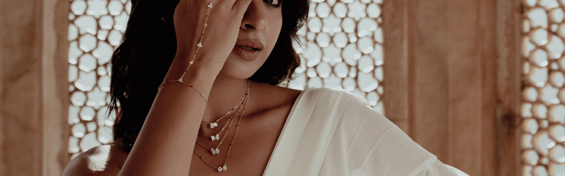 Elegant South Asian woman wearing a layered Modern Mangal mangalsutra with multiple pendants featuring marquise and round-cut diamonds, styled with a dainty gold handchain bracelet. Dressed in a draped white sari and standing against a traditional jaali window in a Jaipur palace, fusing modern mangalsutra design with regal Indian architecture.