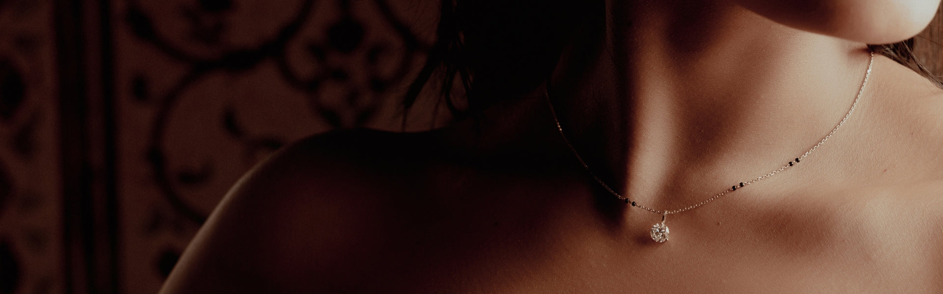 Close-up of a South Asian woman wearing a minimalist Modern Mangal mangalsutra featuring a single round diamond pendant and evenly spaced black beads on a fine gold chain. Captured in low, ambient lighting with an intricately patterned backdrop, the choker-style necklace reflects a modern interpretation of traditional symbolism.
