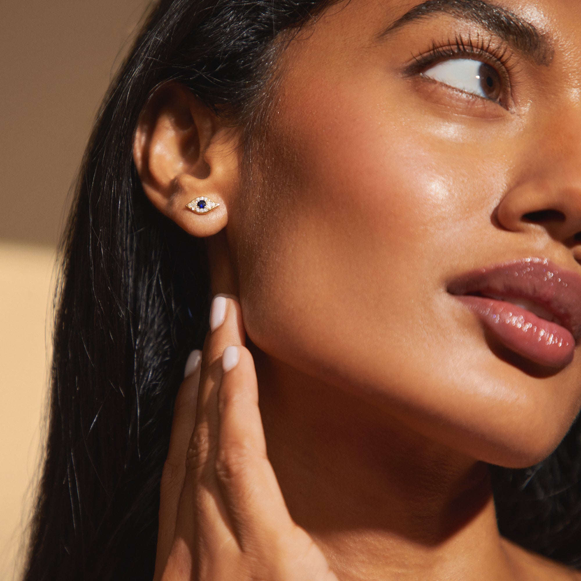Close-up view of model wearing the Zuri Studs in yellow gold, capturing the detailed brilliance of the sapphire and diamond evil eye design nestled against the earlobe.